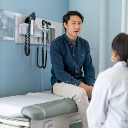 A Korean man is at a routine medical appointment. His doctor is a black woman. The patient is sitting on an examination table facing his doctor.  The kind doctor is listening as he speaks.